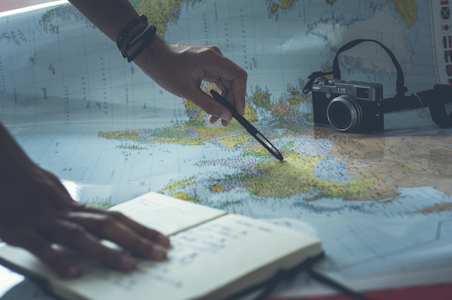 A user checking different locations on a physical world map with the aid of a pen, book, and digital camera.
