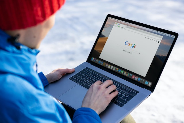 Person in a red knit hat and blue jacket using a MacBook Pro outside to perform a Google search.