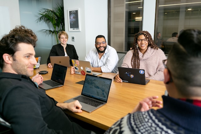 Four people all on laptops, two men and two women, listen to person talking in a board meeting.