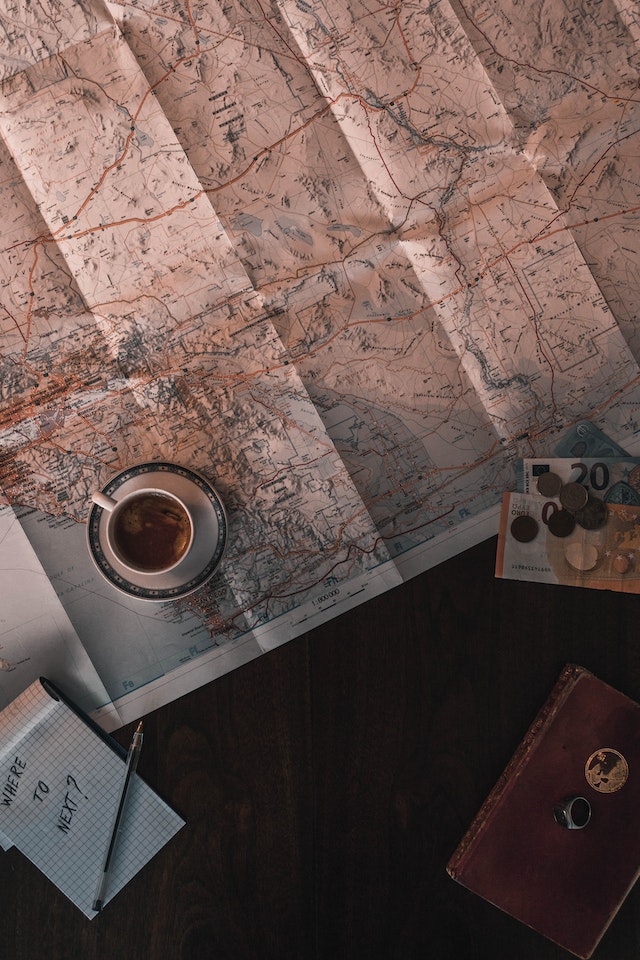 White coffee cup sitting on a map with labeled rivers and mountains next to money and a notepad.