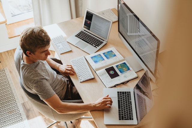 A guy using mapping software on three PCs and a book to visualize his geographic data. 