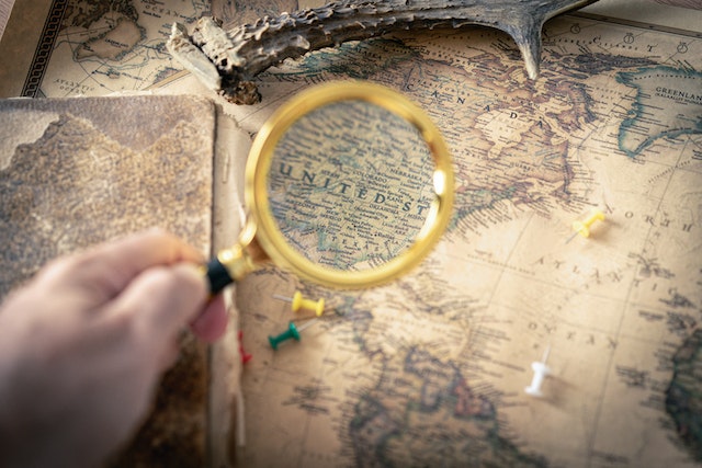 Unseen person holding a magnifying glass over a map of the United States ready to add pins to measure distance.