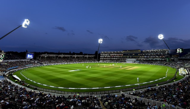 An open-field cricket stadium with thousands of spectators.